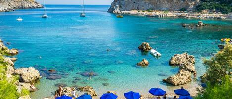 A coastal beach with clear turquoise water, rocky formations, and a sandy area with blue umbrellas.
