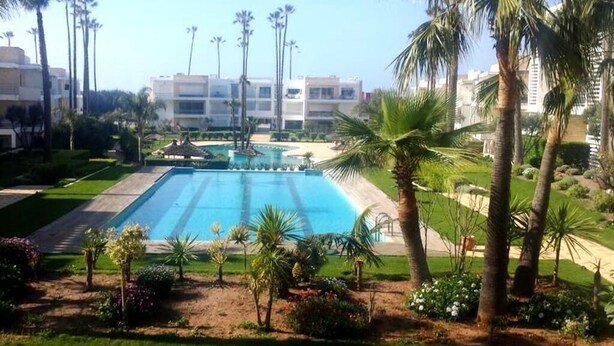 A swimming pool surrounded by palm trees and greenery, with a building in the background. - Very Nice apartment in Eden Tamaris 4175 (Dar Bouazza)