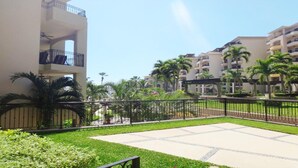 Un patio con suelo empedrado, rodeado de vegetación y palmeras, con edificios de varios pisos al fondo.