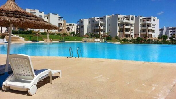 Une piscine entourée de chaises longues et dotée d’un parasol en chaume, le tout bordé d’immeubles d’appartements modernes.