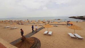 Une plage bordée de rangées de chaises longues et de parasols, une promenade en bois, et quelques personnes qui se promènent.