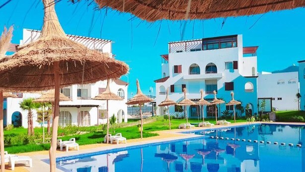 A swimming pool with lounge chairs and thatched umbrellas, surrounded by modern buildings with balconies. - Beautiful Cabo pearl apartment 4442 (Cabo Negro)
