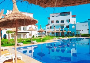 A swimming pool with lounge chairs and thatched umbrellas, surrounded by modern buildings with balconies. - Beautiful Cabo pearl apartment 4442 (Cabo Negro)