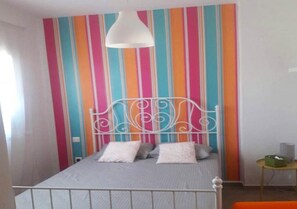Uma cama com estrutura de metal e roupa de cama branca, parede atrás com padrão listrado colorido e luminária pendente branca acima.