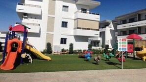 Un terrain de jeux avec des toboggans et des balançoires, situé devant un bâtiment moderne doté de nombreux balcons.