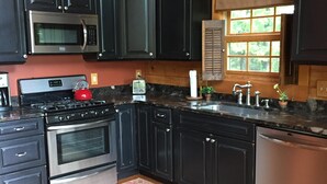 A modern kitchen with dark cabinetry, stainless steel appliances, a wooden floor, and a window with blinds.