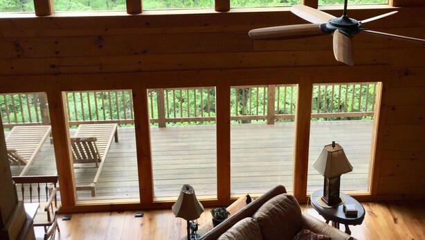 A cabin with large windows offering a view of a forested area. There is a ceiling fan, a couch, and a table with a lamp.