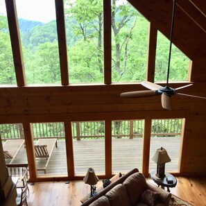 A cabin with large windows offering a view of a forested area. There is a ceiling fan, a couch, and a table with a lamp. - Log Cabin on 9 private acres on Blue Ridge Parkway with gorgeous views (Laurel Springs)