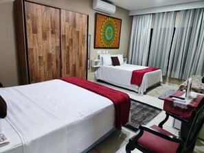 A hotel room with two beds, a desk with a chair, a large window with curtains, and a wall-mounted air conditioner.