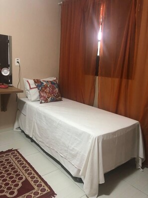 Un lit simple avec une couvre-lit blanche, un oreiller à motifs floraux et un rideau marron partiellement tiré.