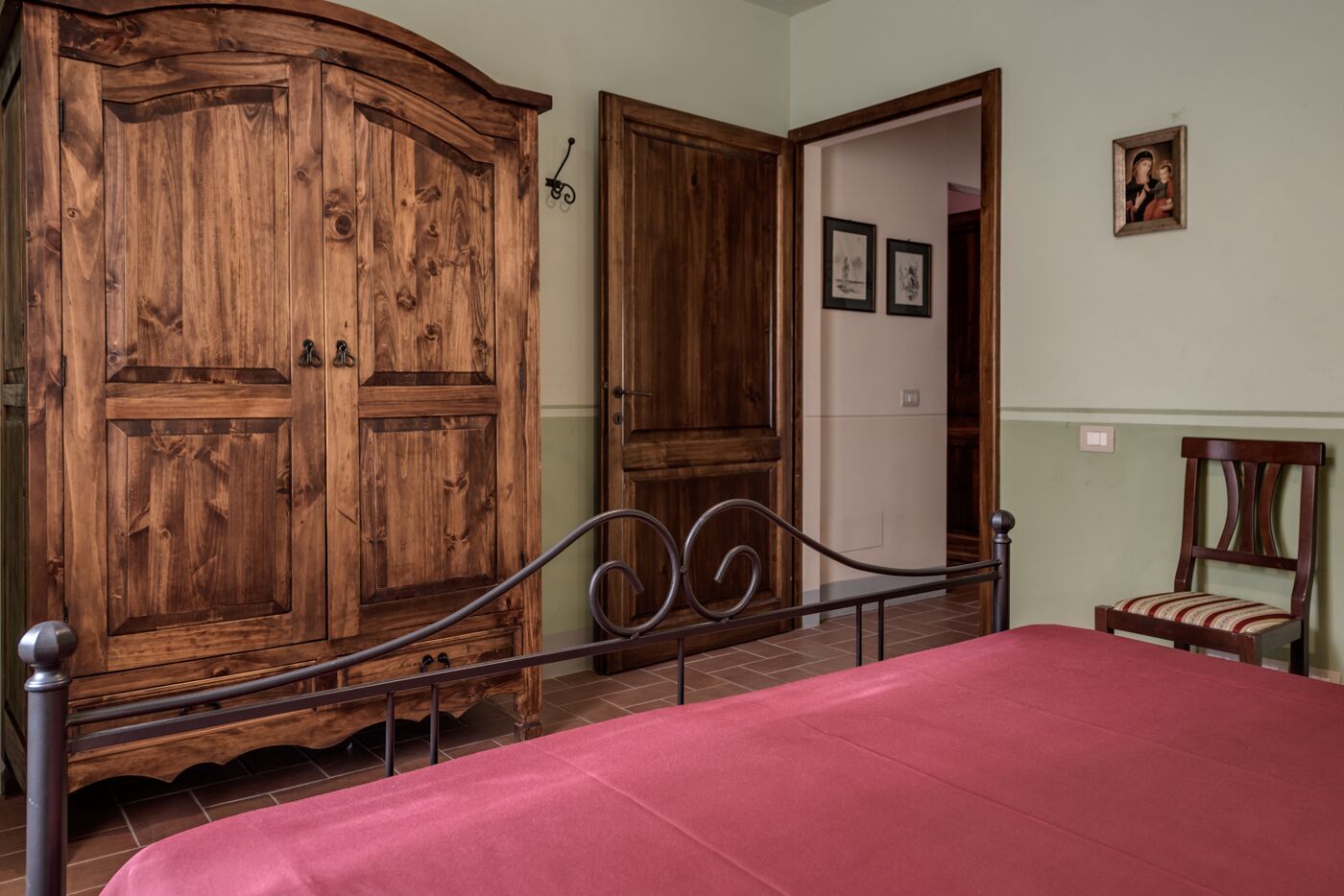 Une chambre avec une grande armoire en bois, un lit recouvert d’une couverture rouge, une chaise et deux tableaux encadrés accrochés au mur.