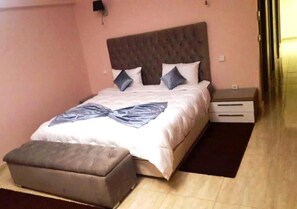 Quarto com cama, mesa de cabeceira, aparador e um banquinho.