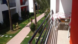 A balcony with a white railing, a small table with a floral tablecloth, and a view of a building with red accents and a pathway leading to it. - Apartment foot in the water in Sary beach 3958 (Sidi Rahal)
