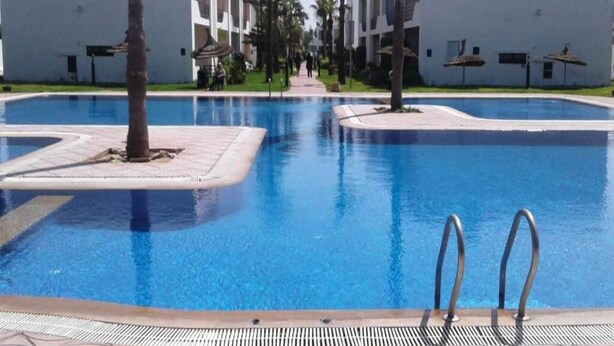 A swimming pool with two palm trees and a white building in the background. - Apartment foot in the water in Sary beach 3958 (Sidi Rahal)