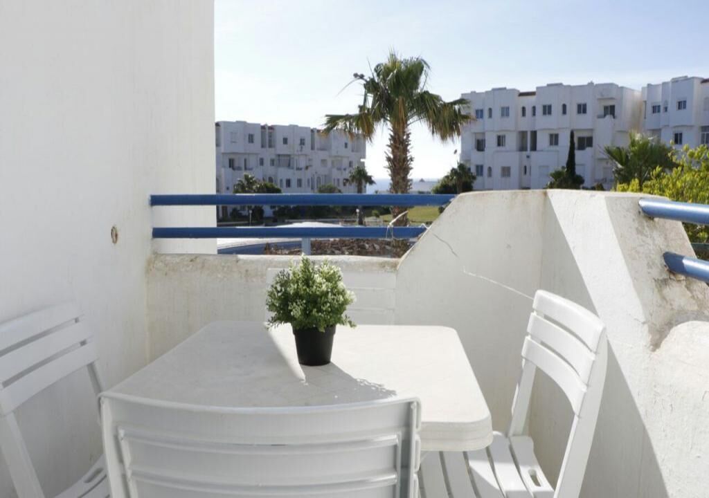 A balcony with a table and chairs, a potted plant, and a view of white buildings and palm trees.