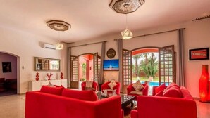 A spacious living room with red sofas, a coffee table, and a TV. There are decorative items on a console table and a large window with a view of a pool.