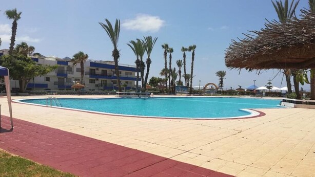 Una piscina con borde rojo, rodeada de palmeras y un edificio con paredes exteriores azules y blancas.