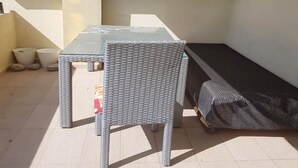 Eine kleine Terrasse im Freien mit Tisch und zwei Stühlen, einer überdachten Couch und einem Fliesenboden.