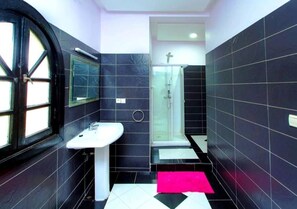 Ein Badezimmer mit schwarzen Fliesen, einem weißen Waschbecken, einer Duschkabine aus Glas und einer rosa Fußmatte.