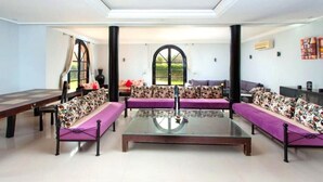 Ein geräumiges Wohnzimmer mit einem Glastisch, zwei Sofas mit gemusterten Kissen, einem Esstisch und einem großen Fenster mit Blick auf Grünflächen.