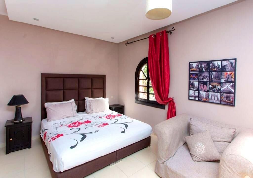 A bedroom with a bed, a nightstand, a chair, a window with red curtains, and a wall-mounted photo collage.