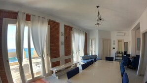 Un vaste espace dédié à la salle à manger, avec une grande table et des chaises bleues, un canapé et une vue sur la mer grâce à de grandes fenêtres.