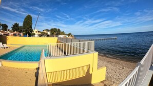 Une piscine avec vue sur la mer, un bâtiment jaune et une jetée qui s’avance dans l’eau.
