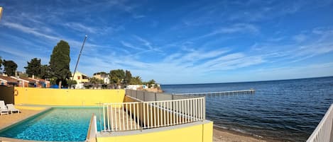 A pool area with a view of the sea, a yellow building, and a pier extending into the water.