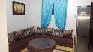 A living room with a sectional sofa, a round table, a refrigerator, and a painting on the wall. - Apartment 5 minutes from the beach 4253 (Saidia)