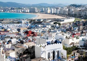 Paisagem costeira urbana com uma praia, edifícios densos e uma estrutura branca imponente com janelas em arco.