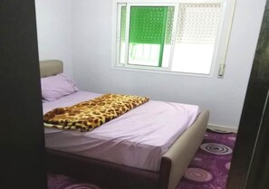 A bedroom with a bed, a window with a green blind, and a purple carpet.