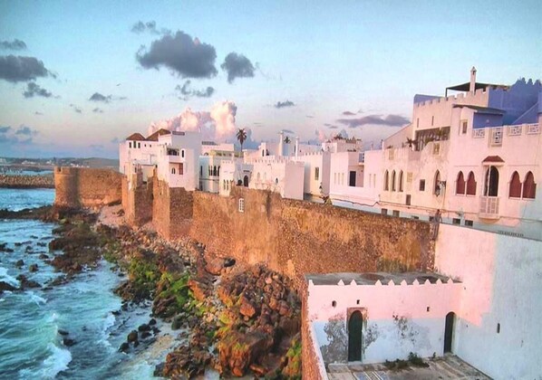 Um edifício costeiro com uma muralha fortificada, uma série de arcos e vista para o mar.