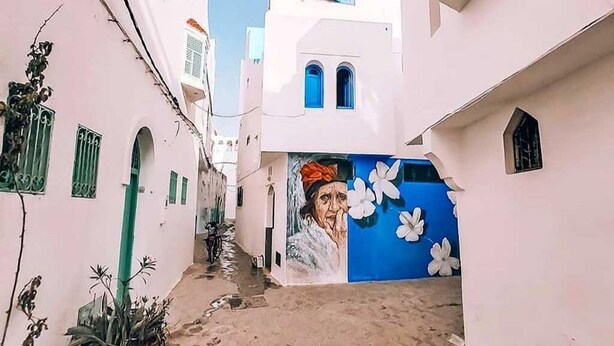 Un estrecho callejón con edificios blancos, contraventanas azules y un mural en la pared.