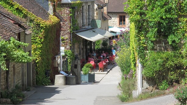 Une rue étroite, bordée de bâtiments des deux côtés, dont certains sont recouverts de lierre. On y voit des tables et des chaises à l’extérieur, ce qui laisse supposer qu’il s’agit d’un café ou d’un restaurant.