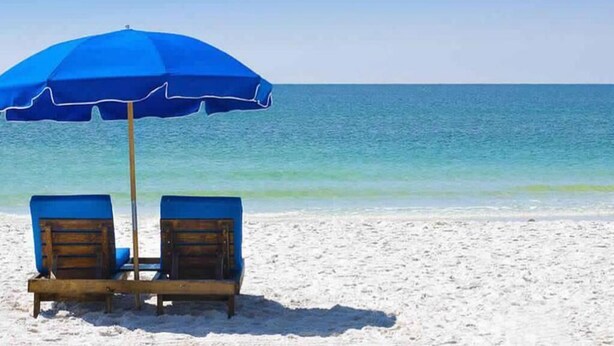 A beach scene with a blue umbrella and two wooden lounge chairs.