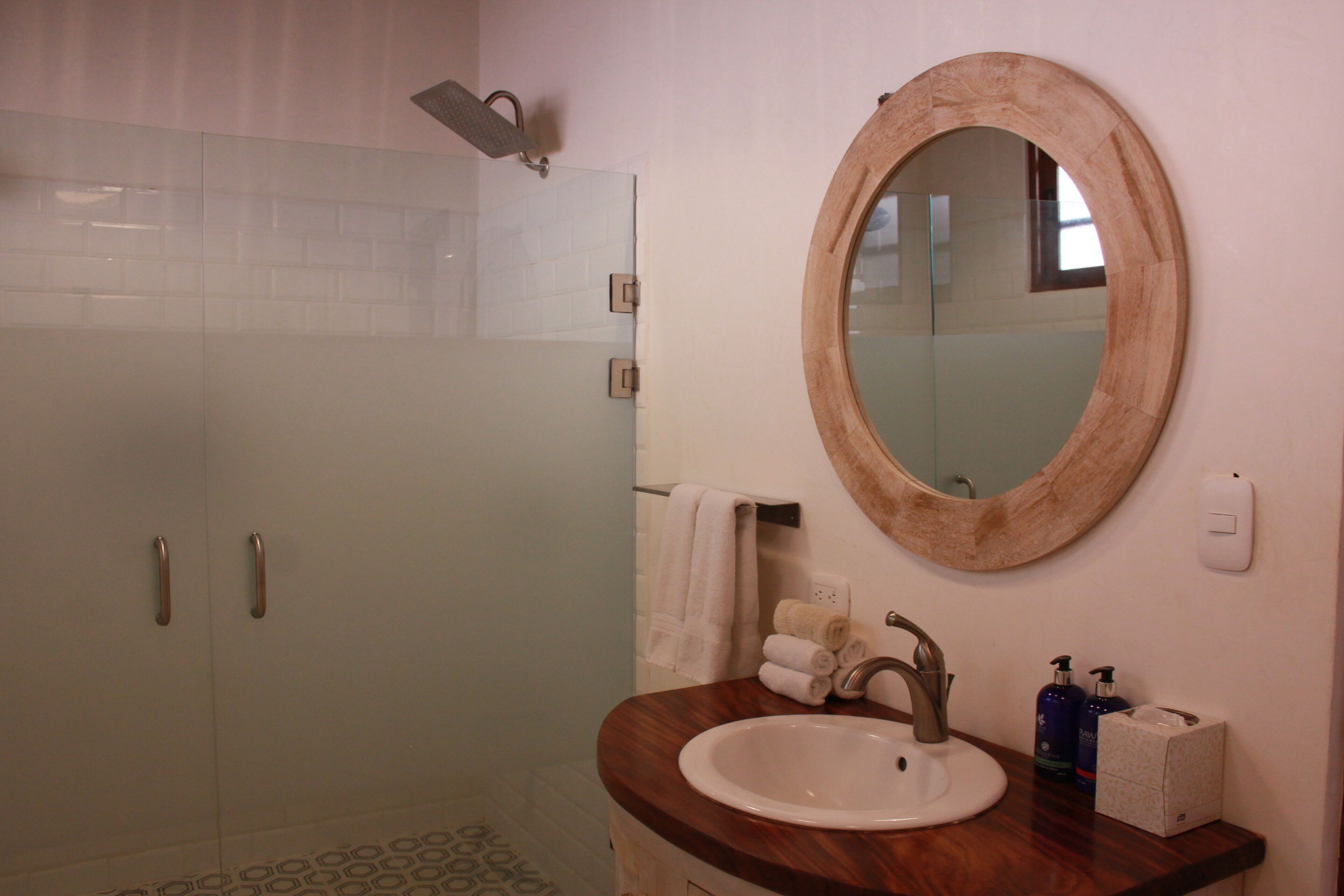 A bathroom with a glass-enclosed shower, a wooden vanity with a sink, and a round mirror.