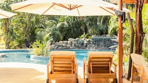 A poolside area with wooden lounge chairs and umbrellas, surrounded by lush greenery and a waterfall feature.