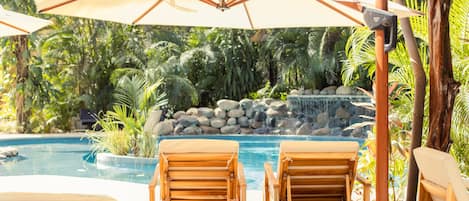 A poolside area with wooden lounge chairs and umbrellas, surrounded by lush greenery and a waterfall feature.