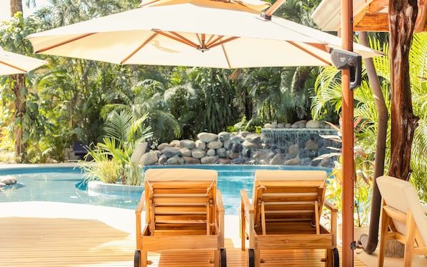 A poolside area with wooden lounge chairs and umbrellas, surrounded by lush greenery and a waterfall feature. - Hotel Guiones (Nosara)