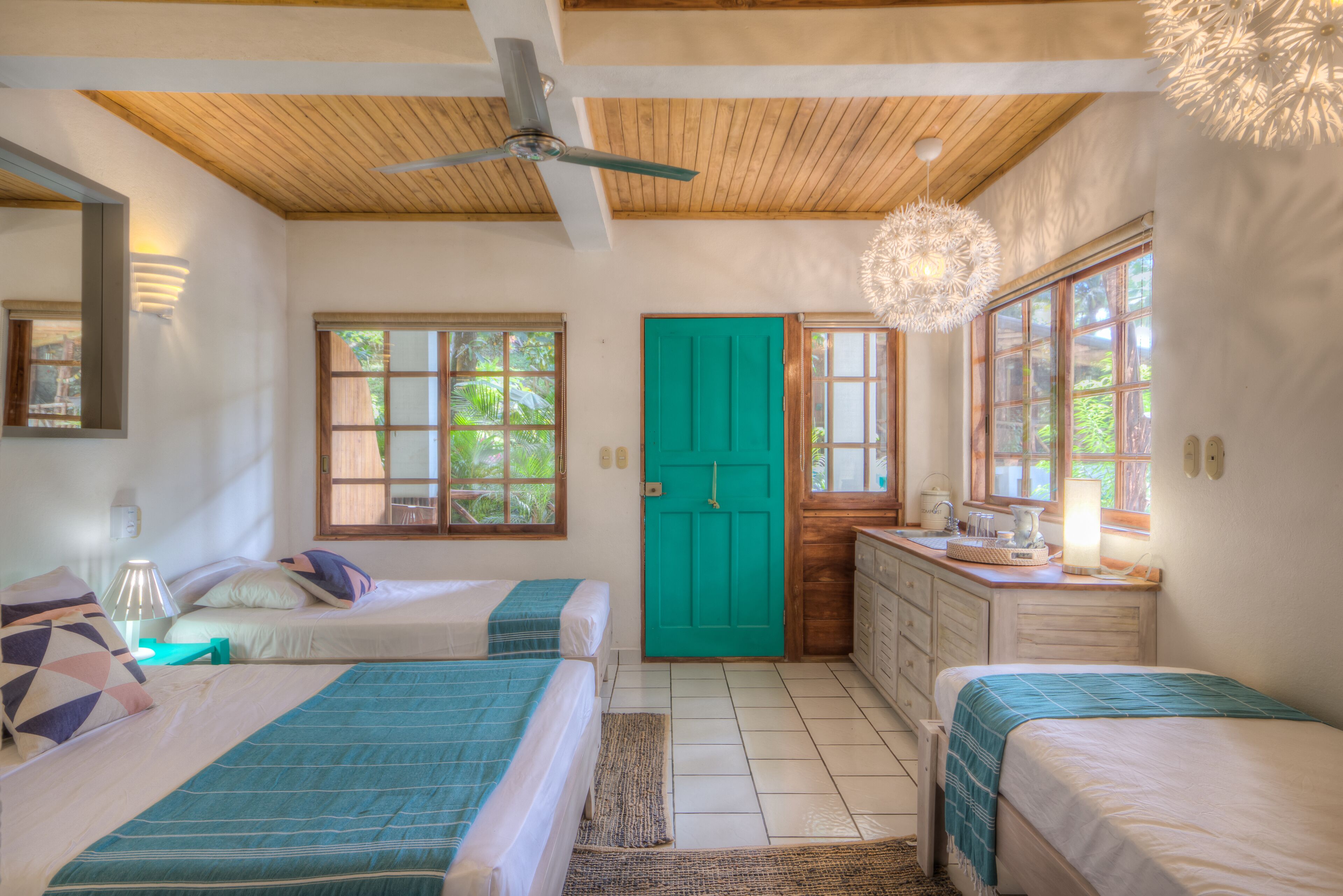 A bedroom with two beds, a wooden ceiling, a green door, and a window with a view of greenery.