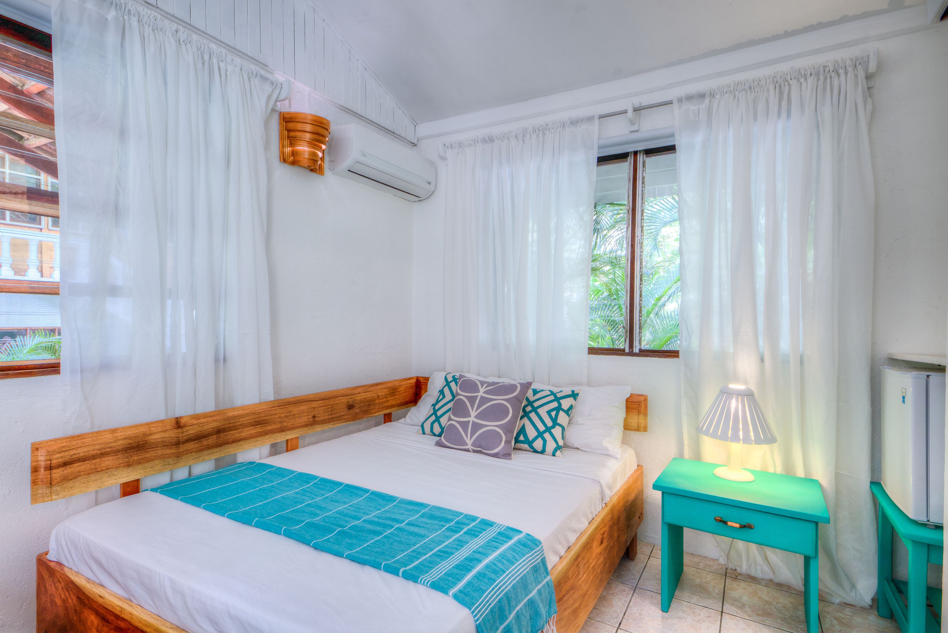 Una camera da letto con un letto di legno, tende bianche, un comodino color acquamarina e una finestra vista sul verde.