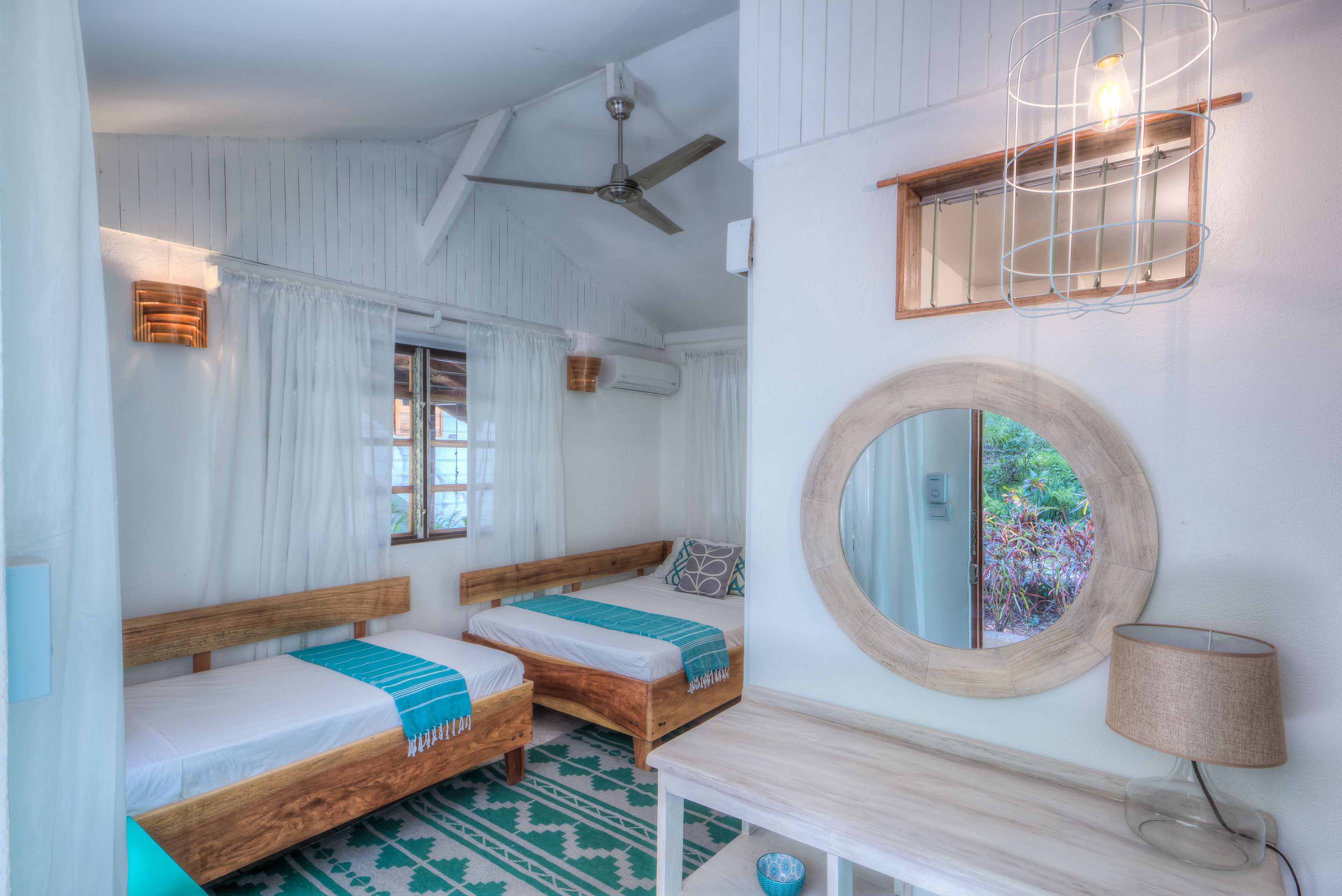A room with two wooden beds, a ceiling fan, a mirror, and a small table with a lamp.