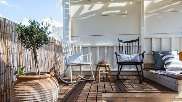 Een terras buiten met een houten terras, witte schommelstoelen, een tafeltje en een potplant.
