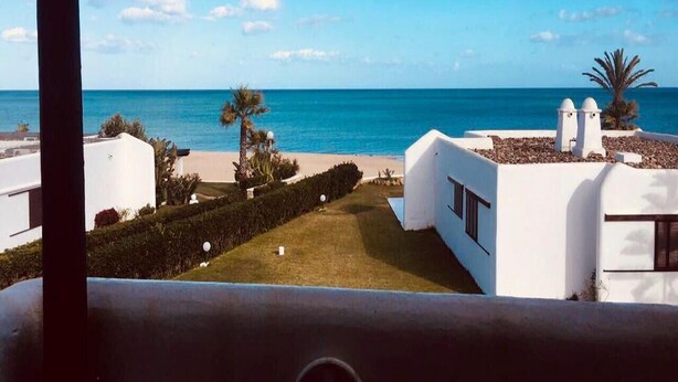 A coastal view with a clear sky, a beach, and modern white buildings with a well-maintained lawn and palm trees. - Beachfront villa in Mdiq 4289 (Mdiq)