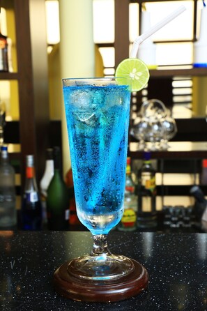 A tall glass with a blue beverage, a lime slice, and a straw, placed on a wooden coaster. - Tolip Olympia Hotel (Addis Ababa)