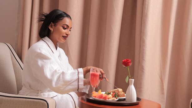 A person sitting on a chair with a tray containing food and a drink, a rose in a vase, and curtains in the background. - Tolip Olympia Hotel (Addis Ababa)