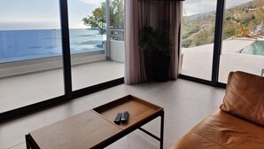 A modern living room with a leather sofa, a wooden coffee table, and a large window offering a view of the ocean and a hillside. - Private swimming pool, amazing views, sleeps 6. (Rethymno)