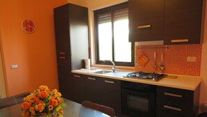 Une cuisine avec des meubles de couleur sombre, un plan de travail blanc, une cuisinière à gaz et une fenêtre avec des stores.