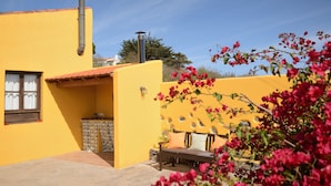 A yellow building with a chimney, a window with curtains, a bench, and a small table with a plant.