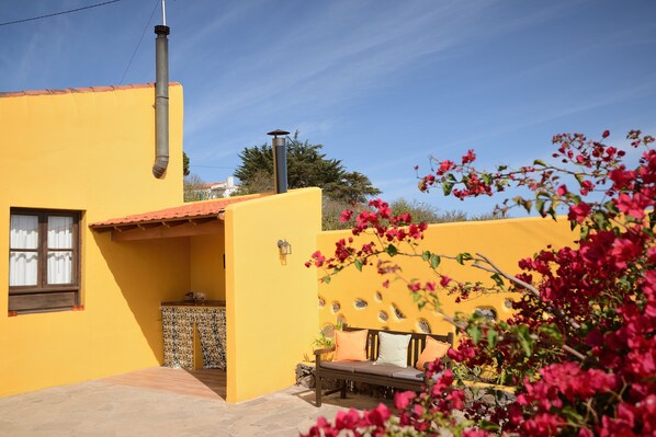 Un edificio amarillo con chimenea, una ventana con cortinas, un banco y una pequeña mesa con una planta.